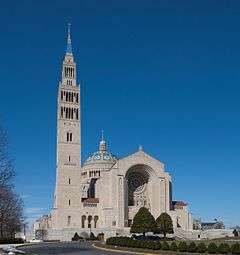 A road with trees in front of the main facade of the basilica, showing the large rose window above the entrance, a dome on top, and a tall bell tower attached to the left of the basilica