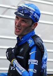 A man in his early thirties, wearing a black and white cycling jersey with blue trim. He also wears a matching cap, with a pair of sunglasses on the cap