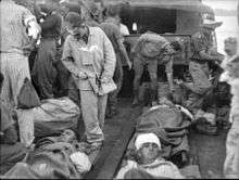 Wounded men lie on stretchers on the deck of a landing craft covered by blankets. Others are tending the wounded.
