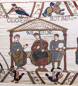 Tapestry image of a seated man in robes holding a sword with WILLELM written above his head flanked by two seated men in chainmail.