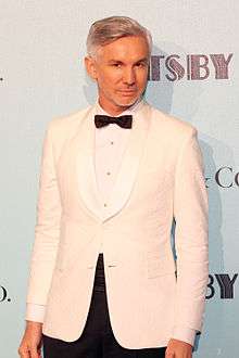 A male with grey hair is seen standing in front of a white wall with black text. He is wearing a white jacket on top of a white shirt with a black bow tie shirt.