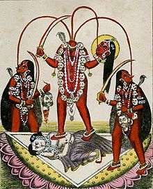 A decapitated, nude, red-complexioned goddess stands on a copulating couple inside a large lotus. She holds her severed head and a scimitar. Three streams of blood from her neck feed her head and two nude, red-coloured women holding a scimitar and a male severed head, who flank her. All three wears a garland of severed heads, a serpent (around her neck) and various gold and pearl jewellery.