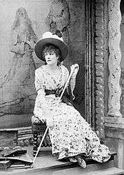 Full length monochrome photograph of a woman seated on a low stool in front of a painting and facing slightly to her right. She wears a high-waisted flowered dress with a solid colour sash and a broad-brimmed hat decorated with flowers and holds a long walking stick.