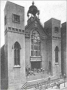 A black and white picture shows the front a three-story building. Two rectangular towers are visible, one on each side of a recessed bay. The towers have pointed arched windows on the bottom and square ones on top. The bay has s dark entrance at the bottom and a sign with Hebrew writing on top of it, surmounted by large arched rose window. Atop the roof of the bay is a four-sided cupola supported by upright beams. Stairs lead from the sidewalk to the entrance, and people are visible standing on the sidewalk and stairs.