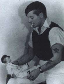 Three-quarter length portrait of a tattooed, short-haired woman in casual, male clothing. She is smoking and looking down at a doll she is holding.