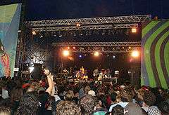 Six musicians playing instruments on a brightly lit stage as a crowd watches.