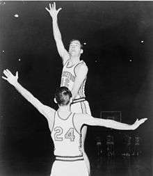 A basketball player, with the number 42 on his short-sleeved uniform, leaping high into the air with his right hand stretched upward.  He is being guarded by another player with the number 24 on his shirt