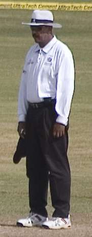 A dark skinned man standing, wearing white shirt, shoes and hat, along with a black pant and sunglasses. A cricket field and the boundary ropes can be seen in the background.