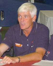 An older, tan Caucasian man with white hair is sitting down on a chair behind a table while speaking to someone off camera. He is wearing a short-sleeved dark blue polo shirt and a gold and silver watch on his left wrist.