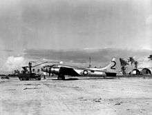 A propeller aircraft sits on a runway. A tracked vehicle with a crane lifts something above it. In the background are a jeep, three Quonset huts and palm trees.