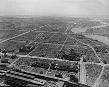 Black and white aerial photo of an urban area comprising several large buildings separated by large fields of rubble. Streets and rivers are visible.