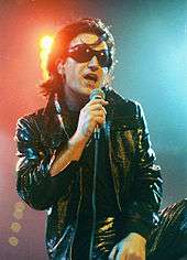 A man with black hair, black sunglasses, and a black leather attire speaks into a microphone.