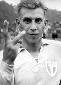 A blonde man raises his right hand to the camera with three fingers up; he is dressed in a light-coloured football kit.