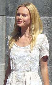 Young blond woman, straight hair below shoulders, wearing a plainly styled, white embossed dress
