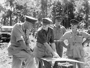 Five soldiers in peaked caps and shirtsleeves study a map on an improvised table in the bush.