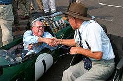 Two elderly men shake hands on a race starting grid. One is sitting in a green racing car, the other in a wheelchair.
