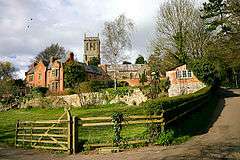 Red brick buildings in front of grey stone church. In the foreground in a grassy field contained within a hedge and fence.