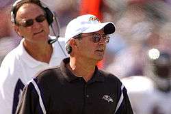 Candid photograph of Zauner standing in front of Brian Billick on a football sideline wearing a black polo shirt and white baseball cap both of which bear the Baltimore Ravens logo