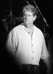 Black and white photograph of Wilson standing onstage looking out to the audience. He is wearing a casual long-sleeved shirt.