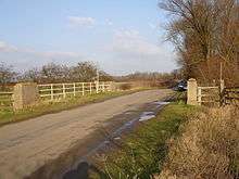 road with concrete pillars and white painted handrails where the drainage ditch passes underneath.  A stand of bare trees on the right and some bare hedgerows on the left show this is winter