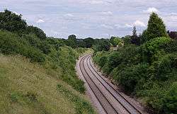 A double-tracked railway line passes through a cutting. The lines are clean and well-maintained, with fresh ballast. The banks of the cutting have light foliage, with few buildings visible nearby.
