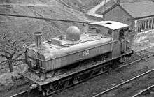 A pannier tank locomotive, seen from above and to the front, is passing through hilly countryside. The locomotive, particularly at the front, is streaked with vertical stains. The lettering "NCB 5774" is shown on the side of the pannier tank.