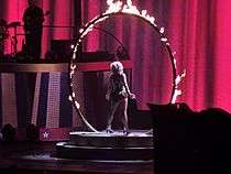 Image of a blond woman. She is standing in the middle of a fiery ring, wearing a black, white and gold outfit, with a pink led screen in front of her. A guitarist can be seen on her left.