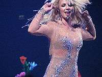 Britney Spears wearing a nude dress with crystals and is holding her arms at the height of her own head.