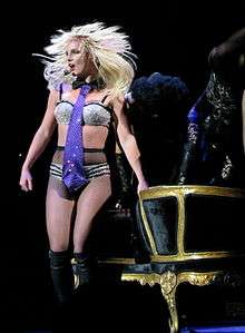 Image of a blond female performer. She has a headset around her hand and is wearing sparkly silver and black lingerie, fishnet stockings and knee-high black boots. She stands in front of a black and golden couch.