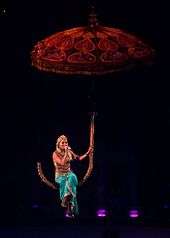 A female blonde performer. She is singing while suspended on a giant umbrella in the air. She wears eastern-inspired clothes.