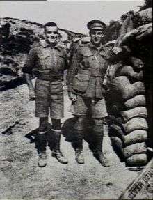Two men in military uniform standing side-by-side. The man on the left is slightly shorter and stockier, while the man on the right has his arm leaning on a stacked up pile of sandbags.