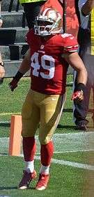 A Caucasian American football player dressed in uniform and protective gear walks on the field.