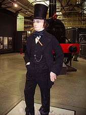 Inside a large building, a life size figure of a man in his late forties, dressed in nineteenth century jacket, trousers and waistcoat, with a prominent watch-chain across his chest. He wears a tall black stove-pipe hat, has long sideburns and has a cigar in his mouth. An old steam locomotive in the background.