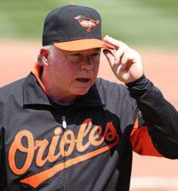 A man wearing a black baseball cap with an orange bill and a bird on the front and a black warm-up jacket with "Orioles" across the chest in orange script touches his left hand to the bill of his cap