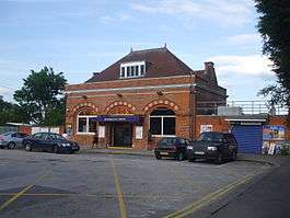 A red-bricked building with a blue sign reading "BUCKHURST HILL STATION" in white letters and four cars parked in the foreground
