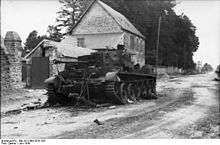 A knocked out tank sits at the side of a road, in front of a two story house.