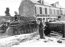 Two tanks are positioned on either side of a street with a soldier standing in the middle of the road; the road is damaged and scattered with debris.