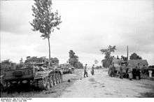 Two tanks are parked on a grass verge to the left of a road while another tank is parked on the right side being inspected by a group of men. Two men are standing in the middle of the road.