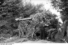 Three men conduct maintenance on a tank; the tank is partially obscured by bushes, which its gun barrel protrudes through.