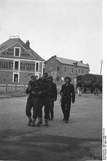 Two wounded Commandos escorted by two armed German naval personnel. A large building is in the background