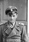 A man wearing a military uniform, peaked cap and a neck order in the shape of a cross. His cap has an emblem in shape of a human skull and crossed bones.