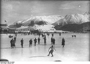 A figure skater performing on a large frozen outdoor area with a group of people nearby on the ice. The background shows snow covered mountains and a building.