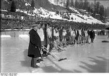 Ice hockey players and staff from two different teams stand together side-by-side in an outdoor ice rink stadium.
