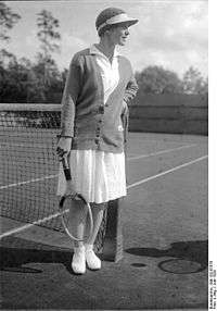 A woman looking away from the camera with a tennis racket in her right hand and a colored sweater on and all white clothing, which this picture is a black and white