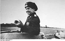 A smiling Munzel wearing a black military uniform stands inside his tank turret.