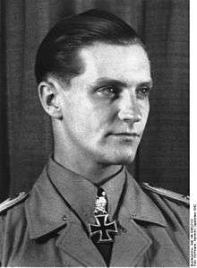 The head and shoulders of a young man, shown in semi-profile. He wears a military uniform with an Iron Cross displayed at the front of his white shirt collar. His hair appears blond and short and combed back, his nose is long and straight, and his facial expression is determined; looking to the left of the camera.