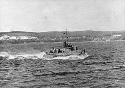 Boat travelling through choppy water leaving a large wake behind it. Cliffs and forested hills can be seen on the shoreline in the background.