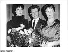 A man in a suit stands in the middle of two formally dressed women, each holding a flower bouquet.