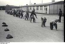 A line of half-naked prisoners performing "leap frog", under supervision of one of the Kapos. In the background the main gate to Mauthausen as well as two wooden barracks are visible.