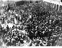 A group several hundred naked men is crowded in an enclosed courtyard, with garage doors visible on three sides.
